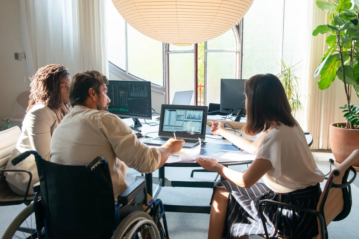 Three colleagues at a meeting table reviewing charts on a laptop and on printed pages